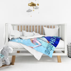 Może przedstawiać: Białe ł&oacute;żeczko dziecięce z niebiesko-białym kocem z motywem księżniczki i imieniem "Sophia". Ł&oacute;żeczko ma drewniane akcenty, a pluszowa zabawka słonia znajduje się w pobliżu. Koc ma wymiary około 150 cm x 200 cm.