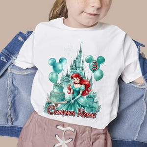 以下が含まれることがあります： 白いTシャツ。赤い髪のプリンセス、お城、風船、ケーキのターコイズ色のイラスト。数字の3と「Custom Name」の文字が表示されています。