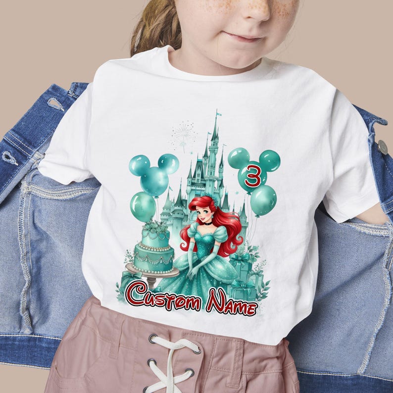 以下が含まれることがあります： 白いTシャツ。赤い髪のプリンセス、お城、風船、ケーキのターコイズ色のイラスト。数字の3と「Custom Name」の文字が表示されています。