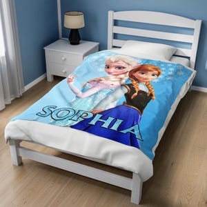 Może przedstawiać: Jasnoniebieski koc z Elsą i Anną z filmu Kraina Lodu. Na kocu znajduje się imię "SOPHIA" wydrukowane dużymi, niebieskimi literami. Koc leży na białej ramie ł&oacute;żka.