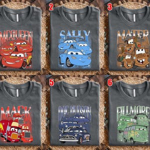 May include: Six gray t-shirts featuring characters from the animated movie "Cars." Each shirt displays a different character's name and image: McQueen, Sally, Mater, Mack, Doc Hudson, and Fillmore. The designs are in various colors.