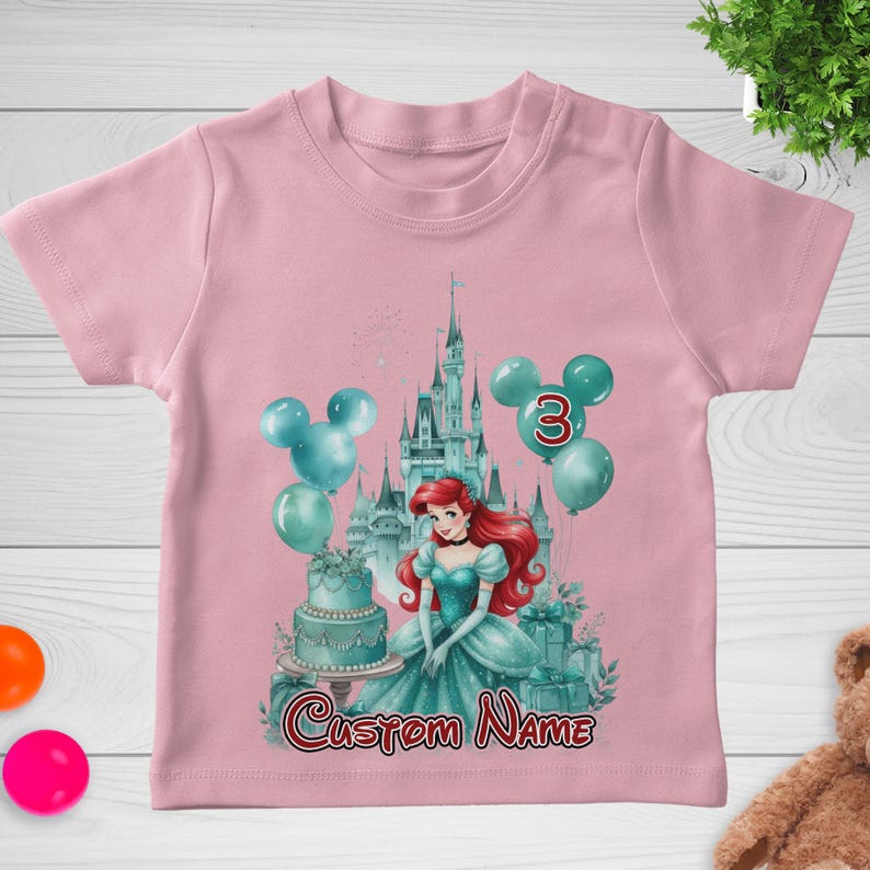以下が含まれることがあります： ピンク色の幼児用Tシャツ。水色のドレスを着たプリンセス、お城、風船、ケーキのグラフィックが特徴です。「3」の数字と「Custom Name」の文字が表示されています。