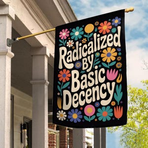May include: Black flag with the words "Radicalized by Basic Decency" in white, surrounded by colorful flowers. The flag is hanging from a gold pole on a porch.