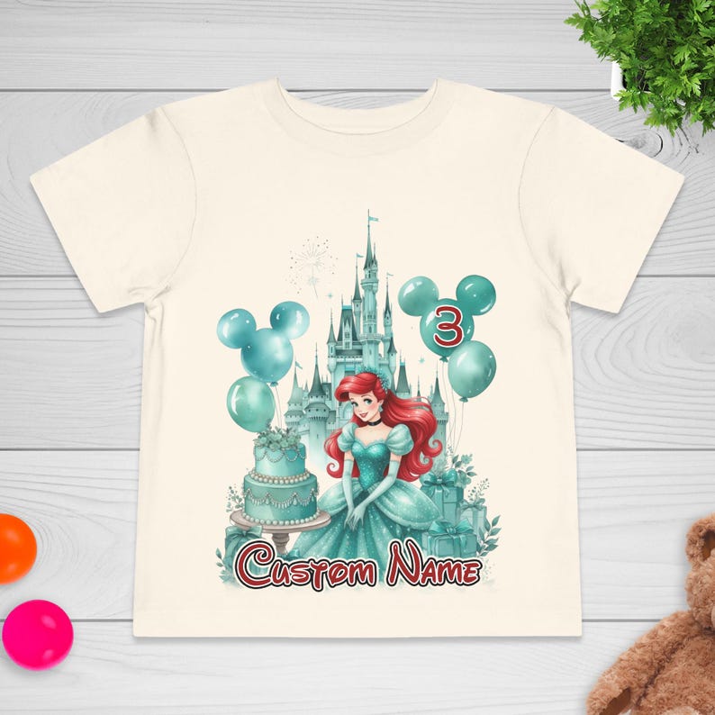 以下が含まれることがあります： クリーム色の幼児用Tシャツ。赤い髪のプリンセス、お城、風船、ケーキのカラフルなグラフィックが特徴です。風船には数字の3が表示され、プリンセスの下には「Custom Name」の文字が書かれています。