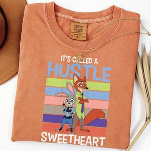 May include: A rust-colored t-shirt featuring a graphic of Judy Hopps and Nick Wilde from Zootopia. The text reads IT'S CALLED A HUSTLE SWEETHEART above and below the characters, set against a background of horizontal stripes in green, blue, and pink.