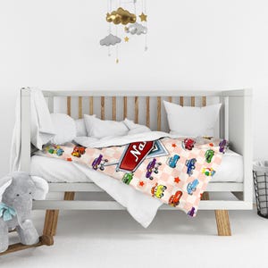May include: A baby blanket with a repeating pattern of colorful cartoon cars on a pink and white checkered background. The blanket is draped over a white crib. A sign with the name "Na" is on the blanket.