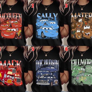 May include: Black t-shirts featuring characters from the animated movie "Cars." Designs include Lightning McQueen, Sally, Mater, Mack, Doc Hudson, and Fillmore. Each shirt has a different graphic and character name.