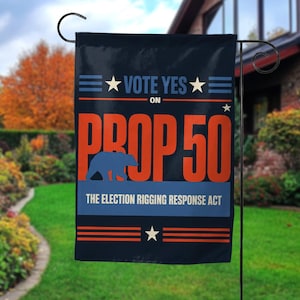 May include: A black garden flag with the text "VOTE YES ON PROP 50" in white and orange. The flag also features a blue bear silhouette and the text "THE ELECTION RIGGING RESPONSE ACT". The flag is hanging in a garden.