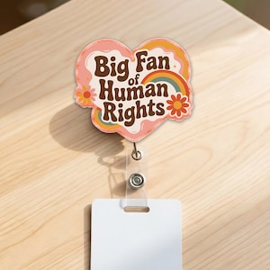 May include: A heart-shaped badge reel with the text "Big Fan of Human Rights" in brown and white. The badge has a retro design with a rainbow and flowers. The reel has a clear plastic strap and a white rectangular card.