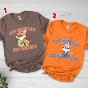 May include: A brown t-shirt featuring a cartoon fox and the text "YOU HUSTLED MY HEART" and an orange t-shirt featuring a cartoon rabbit and the text "YOU ARE MY BEST CATCH". Both shirts have short sleeves and crew necklines with printed graphics.