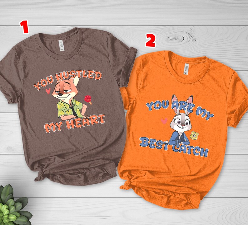May include: A brown t-shirt featuring a cartoon fox and the text "YOU HUSTLED MY HEART" and an orange t-shirt featuring a cartoon rabbit and the text "YOU ARE MY BEST CATCH". Both shirts have short sleeves and crew necklines with printed graphics.