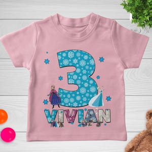 May include: Pink short-sleeved t-shirt featuring a large number 3 filled with snowflakes. The name "VIVIAN" is printed below the number. The shirt also has images of characters from the movie Frozen and snowflake accents.