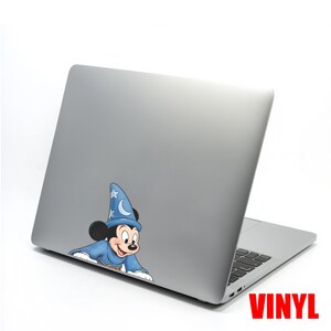 Puede incluir: Un port&aacute;til plateado con una pegatina de vinilo de Mickey Mouse vestido de mago. Mickey lleva una t&uacute;nica y un sombrero azules con estrellas y una luna creciente. La palabra "VINYL" est&aacute; impresa en rojo.