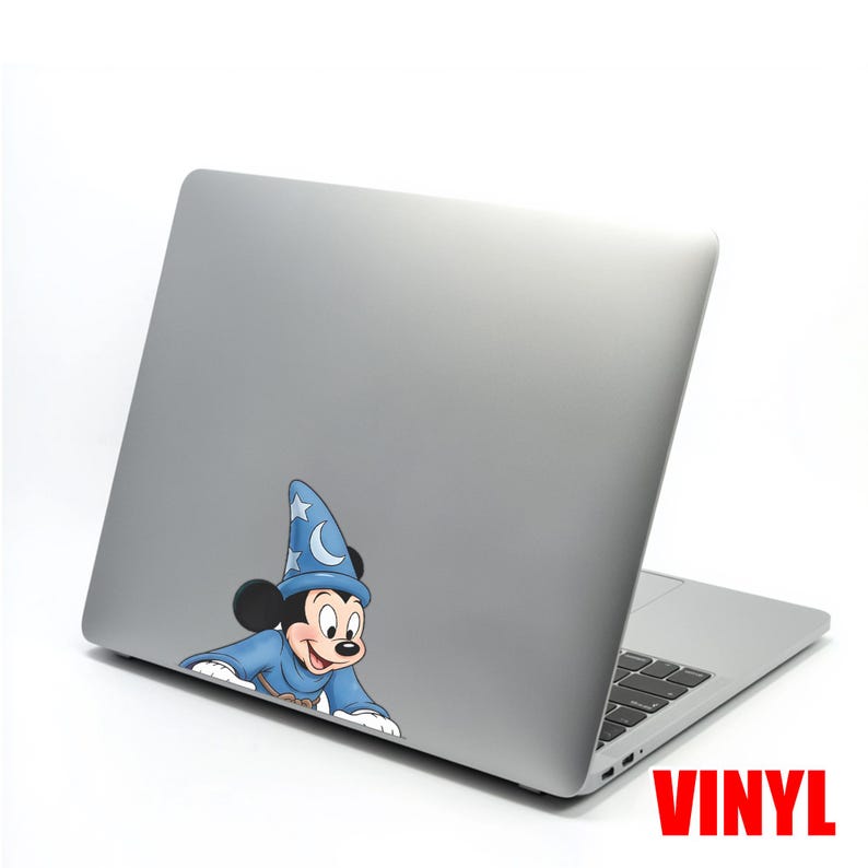 Puede incluir: Un port&aacute;til plateado con una pegatina de vinilo de Mickey Mouse vestido de mago. Mickey lleva una t&uacute;nica y un sombrero azules con estrellas y una luna creciente. La palabra "VINYL" est&aacute; impresa en rojo.