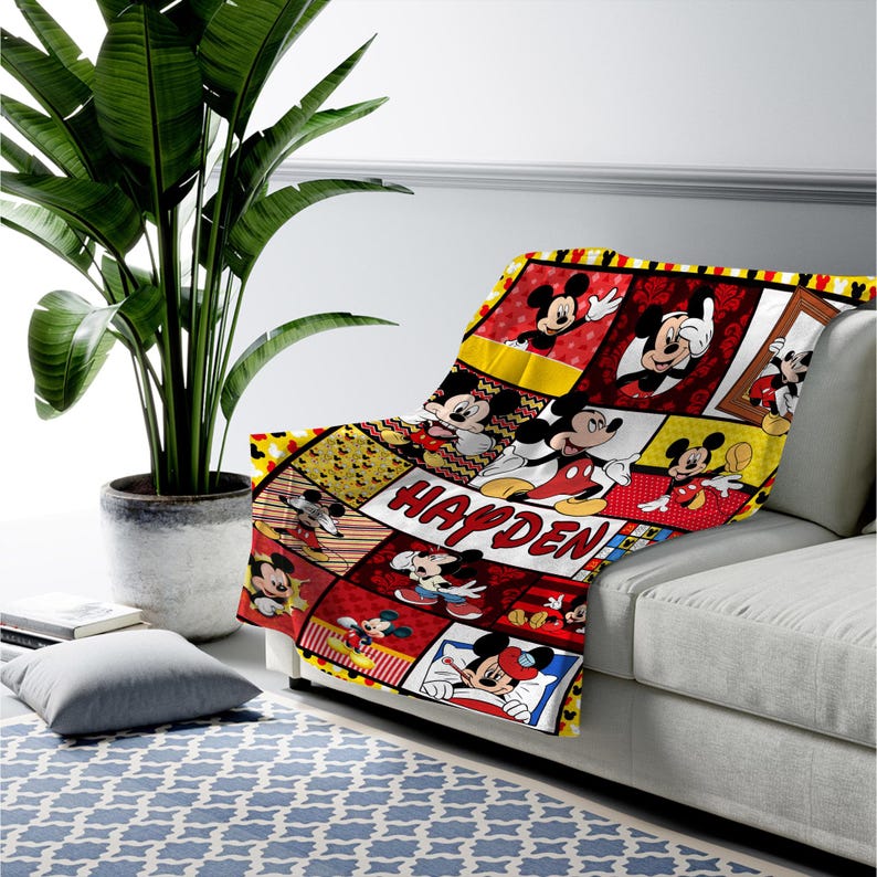 Pu&ograve; includere: Una coperta colorata con un collage di immagini di Topolino in varie pose. La coperta ha uno schema di colori rosso, giallo e nero con il nome "HAYDEN" stampato in bianco. &Egrave; drappeggiata su un divano grigio.