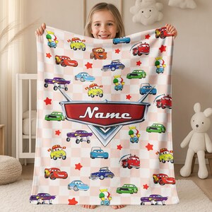 May include: A light pink and white checkered blanket featuring cartoon cars and red stars. The blanket has a large red and silver emblem with the name "Name" printed on it. The blanket is held by a young girl.