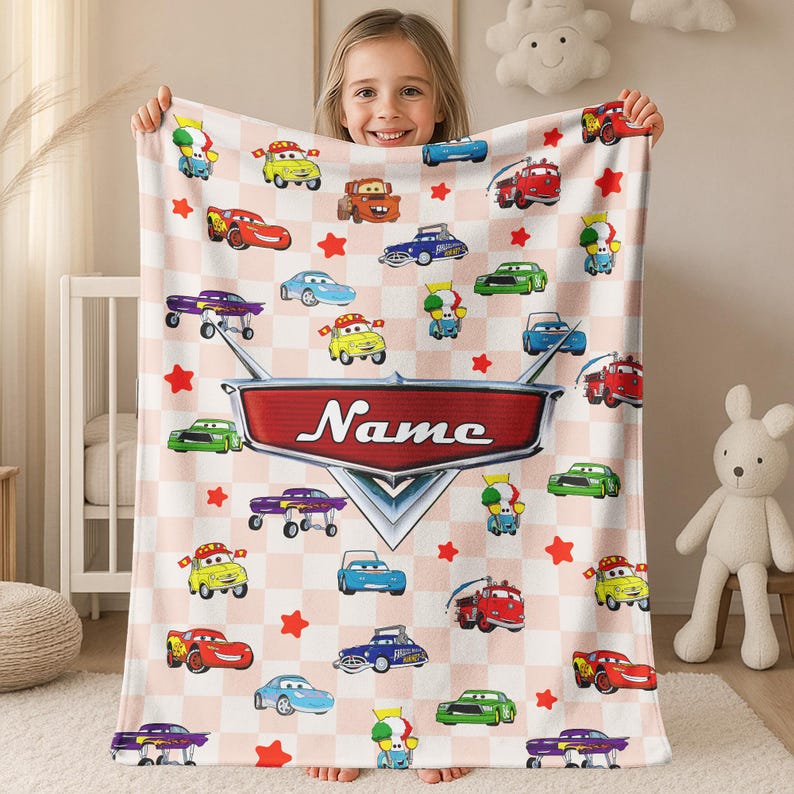 May include: A light pink and white checkered blanket featuring cartoon cars and red stars. The blanket has a large red and silver emblem with the name "Name" printed on it. The blanket is held by a young girl.