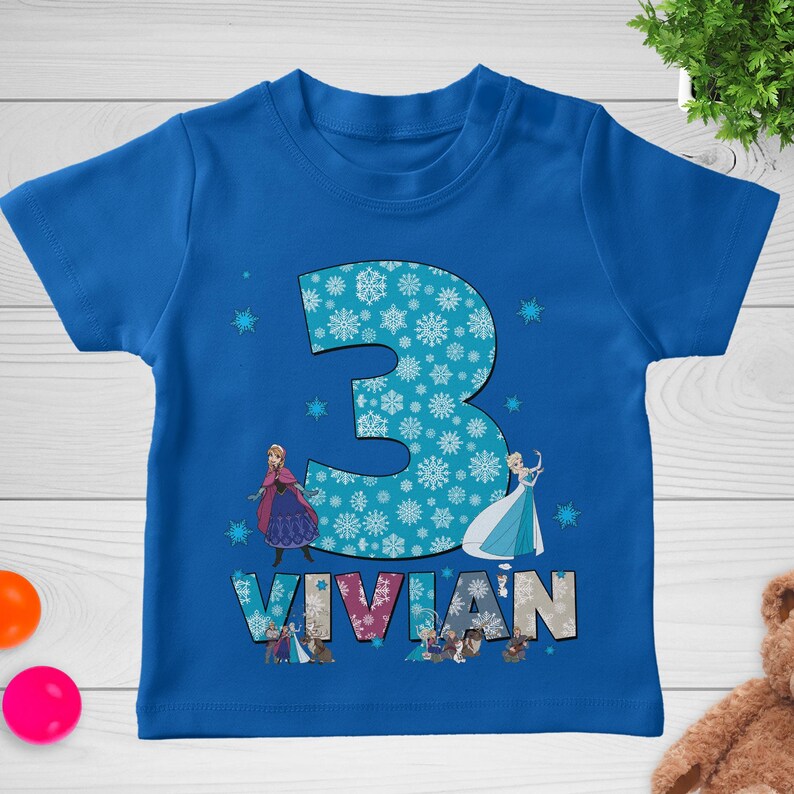 May include: A blue toddler t-shirt featuring the number "3" filled with snowflakes. The name "VIVIAN" is printed below the number, with images of animated characters. The shirt is a vibrant blue.