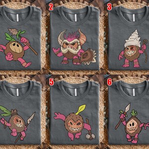 Puede incluir: Seis camisetas grises con dise&ntilde;os de personajes de dibujos animados. Cada camiseta presenta una criatura diferente con acentos rosas, marrones y verdes. Las criaturas est&aacute;n representadas con varios accesorios. Las camisetas se muestran sobre una superficie tejida.
