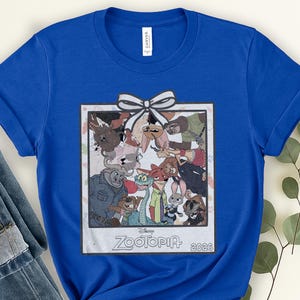 Puede incluir: Camiseta azul real con un gr&aacute;fico de la pel&iacute;cula de Disney Zootopia. El gr&aacute;fico es una imagen enmarcada de los personajes de la pel&iacute;cula con un lazo blanco en la parte superior. El texto "Zootopia 2026" est&aacute; en la parte inferior de la imagen.