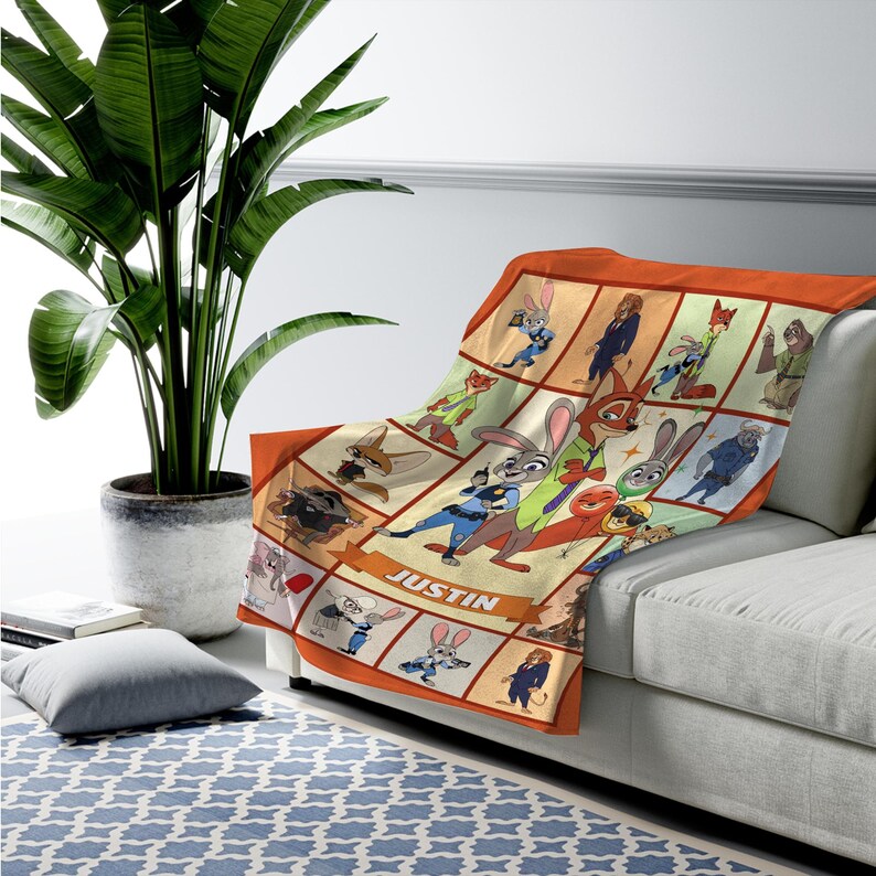 May include: A Zootopia-themed blanket featuring a collage of characters from the animated film. The blanket has an orange border and a cream-colored background with various character illustrations. The name "JUSTIN" is displayed on a banner.