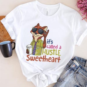 Op de afbeelding: Wit T-shirt met een cartoon vos met een zonnebril en een koffiekopje. Het shirt heeft de tekst "it's called a HUSTLE Sweetheart" in kleurrijke letters. De vos draagt een groen overhemd en een stropdas met patroon.