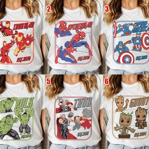 K&ouml;nnte beinhalten: Wei&szlig;e T-Shirts mit Designs von Marvel-Superhelden. Zu den Charakteren geh&ouml;ren Iron Man, Spider-Man, Captain America, Hulk, Thor und Groot. Jedes Design ist mit den Namen der Charaktere und dem Jahr ihres Deb&uuml;ts umrahmt. Eine Auswahl an Kleidung zum Thema Superhelden.