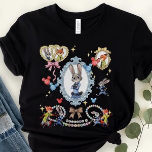 May include: Black t-shirt featuring characters from Zootopia. The design includes Judy Hopps, Nick Wilde, and other characters in oval frames and around the text "WELCOME TO ZOOTOPIA". Decorative elements include bows, balloons, and stars.