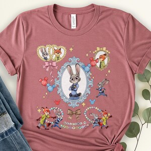 May include: A dusty rose t-shirt featuring cartoon characters from Zootopia. The design includes Judy Hopps, Nick Wilde, and other characters in ornate frames, with the text "WELCOME TO ZOOTOPIA".