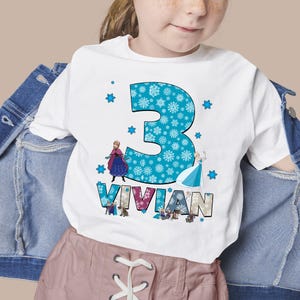 May include: White t-shirt featuring a large blue number 3 filled with snowflakes. The name "VIVIAN" is below the number, with images of animated characters. The shirt is worn by a child.