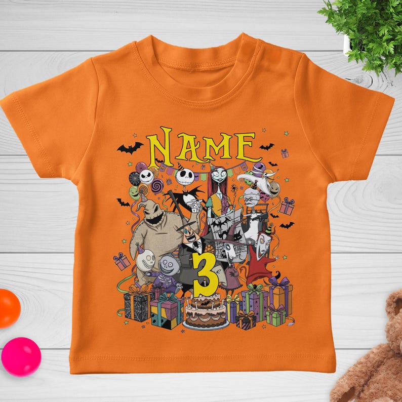 May include: Orange short-sleeved t-shirt featuring a colorful graphic of characters from The Nightmare Before Christmas. The design includes the word "NAME" above the characters and the number "3" in yellow. Presents and a cake are also included in the design.
