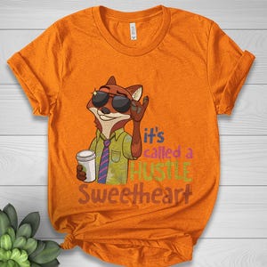 Op de afbeelding: Oranje t-shirt met een cartoon vos met een zonnebril en een koffiekopje. De vos maakt een rock-on handgebaar. De tekst op het shirt luidt "It's called a HUSTLE Sweetheart."