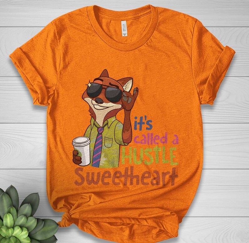 Op de afbeelding: Oranje t-shirt met een cartoon vos met een zonnebril en een koffiekopje. De vos maakt een rock-on handgebaar. De tekst op het shirt luidt "It's called a HUSTLE Sweetheart."