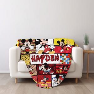 Pu&ograve; includere: Una coperta colorata con un collage di immagini di Topolino in varie pose. La coperta ha uno schema di colori rosso, giallo e nero, con il nome "HAYDEN" ben visibile in una cornice bianca. La coperta &egrave; drappeggiata su un divano bianco.