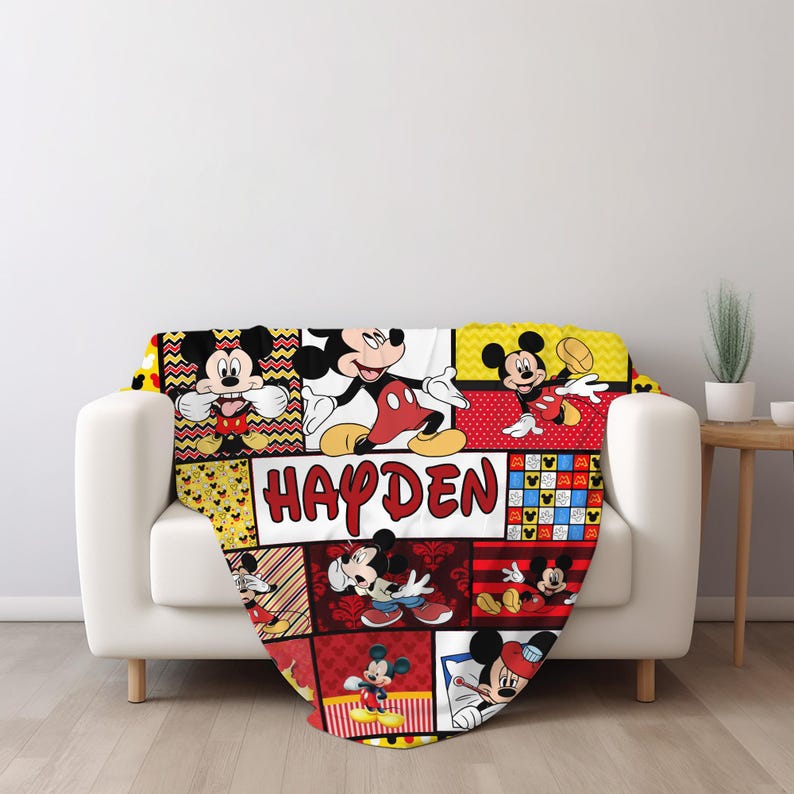Pu&ograve; includere: Una coperta colorata con un collage di immagini di Topolino in varie pose. La coperta ha uno schema di colori rosso, giallo e nero, con il nome "HAYDEN" ben visibile in una cornice bianca. La coperta &egrave; drappeggiata su un divano bianco.