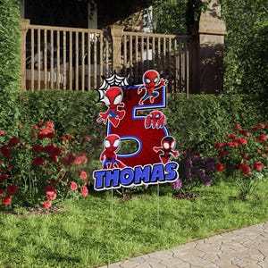May include: A yard sign featuring the number 5 in red with a spiderweb design and several Spider-Man figures. The name "Thomas" is displayed in blue and white text at the bottom. The sign is placed in a grassy yard with flowers.