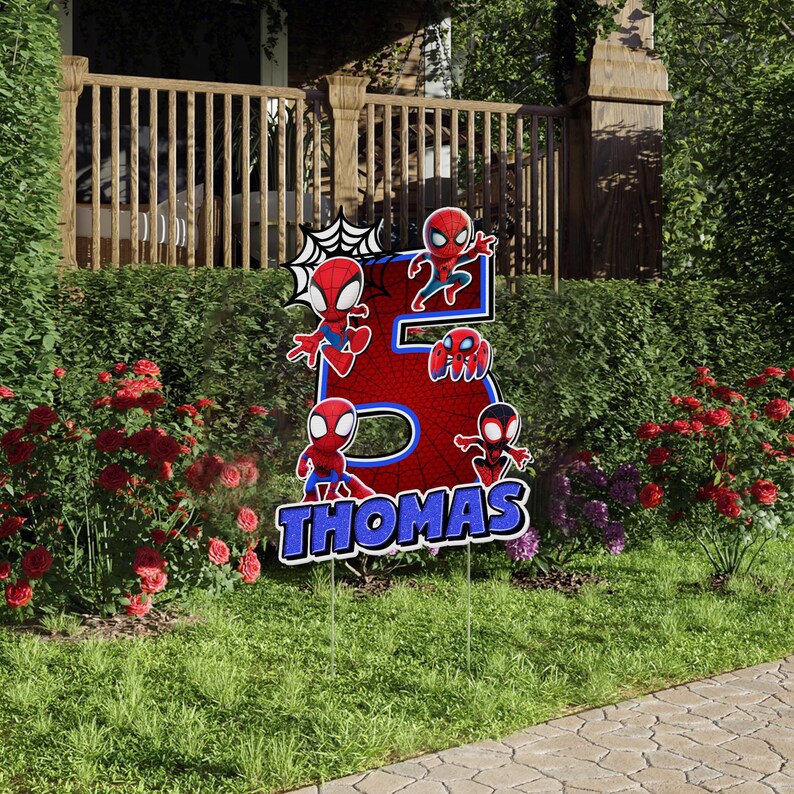 May include: A yard sign featuring the number 5 in red with a spiderweb design and several Spider-Man figures. The name "Thomas" is displayed in blue and white text at the bottom. The sign is placed in a grassy yard with flowers.