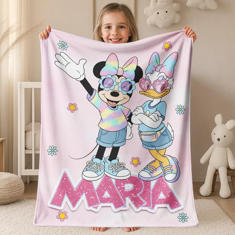 Op de afbeelding: Een roze fleece deken met Minnie Mouse en Daisy Duck in zonnebrillen en tie-dye outfits. De deken heeft de naam "MARIA" in grote roze letters en is versierd met bloemen.