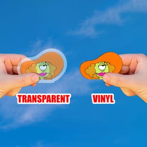 May include: Two stickers, one transparent and one vinyl, featuring a cartoon character with an orange hat and green skin. The word "TRANSPARENT" is printed in red on the left, and "VINYL" is printed on the right.