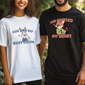 May include: A white t-shirt featuring a cartoon rabbit and the text "YOU ARE MY BEST CATCH" in blue, alongside a black t-shirt featuring a cartoon fox and the text "YOU HUSTLED MY HEART" in orange, both with small red heart graphics.