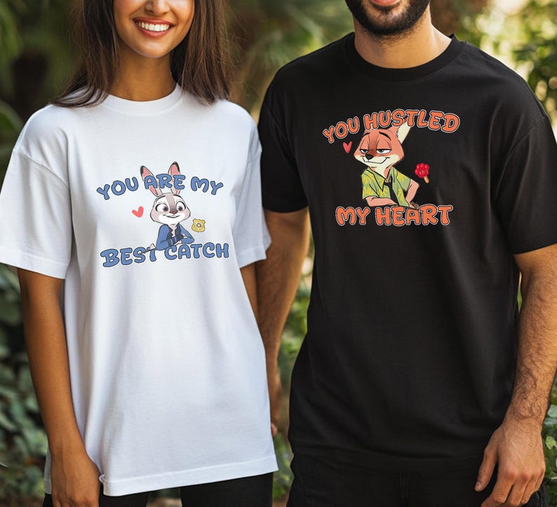 May include: A white t-shirt featuring a cartoon rabbit and the text "YOU ARE MY BEST CATCH" in blue, alongside a black t-shirt featuring a cartoon fox and the text "YOU HUSTLED MY HEART" in orange, both with small red heart graphics.