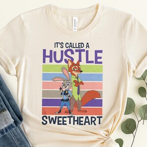 May include: A cream-colored crew neck t-shirt featuring a graphic of a cartoon rabbit and fox. The text reads IT'S CALLED A HUSTLE SWEETHEART. The design includes horizontal stripes in purple, green, blue, pink, brown, and orange behind the characters.