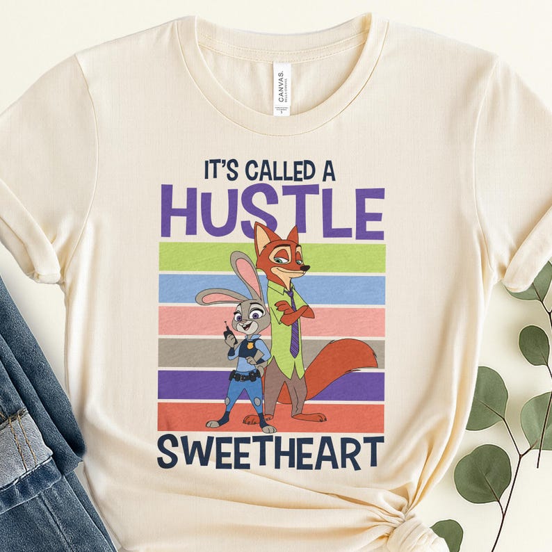 May include: A cream-colored crew neck t-shirt featuring a graphic of a cartoon rabbit and fox. The text reads IT'S CALLED A HUSTLE SWEETHEART. The design includes horizontal stripes in purple, green, blue, pink, brown, and orange behind the characters.