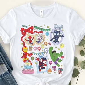 May include: A white short-sleeve t-shirt featuring a colorful graphic print of various superheroes, including Iron Man, Hulk, and Spider-Man characters. The design includes birthday-themed elements like balloons, gifts, a cake, and the text NAME.
