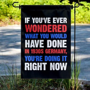May include: Black garden flag with white, red, and blue text. The text reads: "IF YOU'VE EVER WONDERED WHAT YOU WOULD HAVE DONE IN 1930S GERMANY, YOU'RE DOING IT RIGHT NOW."