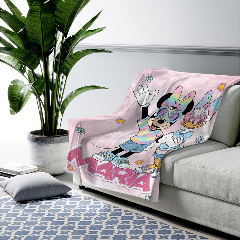 Op de afbeelding: Een lichtroze deken met Minnie Mouse en Daisy Duck met zonnebrillen en kleurrijke outfits. De deken heeft het woord "MARA" in roze letters en is versierd met kleine bloemmotieven. De deken is over een grijze bank gedrapeerd.