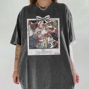 Puede incluir: Camiseta gris oscuro con un gr&aacute;fico colorido de personajes de la pel&iacute;cula de Disney Zootopia. El gr&aacute;fico est&aacute; enmarcado con un borde blanco y un lazo blanco en la parte superior. El texto "Zootopia 2023" est&aacute; impreso en la parte inferior.