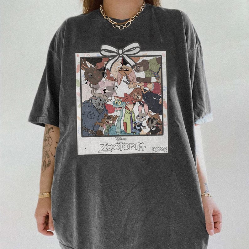 Puede incluir: Camiseta gris oscuro con un gr&aacute;fico colorido de personajes de la pel&iacute;cula de Disney Zootopia. El gr&aacute;fico est&aacute; enmarcado con un borde blanco y un lazo blanco en la parte superior. El texto "Zootopia 2023" est&aacute; impreso en la parte inferior.
