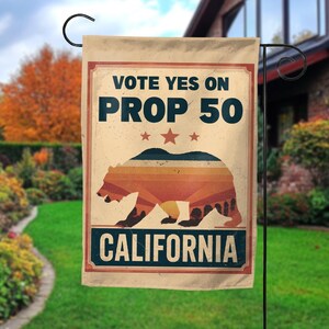 May include: A beige garden flag with the text "VOTE YES ON PROP 50" above a bear silhouette with a striped design. The word "CALIFORNIA" is at the bottom. The flag is hanging on a black metal stand in a grassy yard.