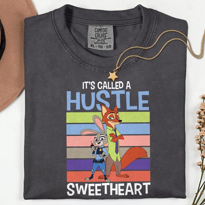 May include: A charcoal gray t-shirt featuring a graphic of a rabbit and a fox character. The text reads IT'S CALLED A HUSTLE SWEETHEART above and below the characters, set against a background of horizontal stripes in green, blue, pink, and brown.